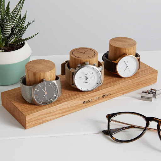 Watch display holder rests on a table, made of a long, wooden block base with 3 thick round columns sticking up enough for a watch band to sit sideways. Customizable text etched into the block base. Watch dial etched into top of the middle column.